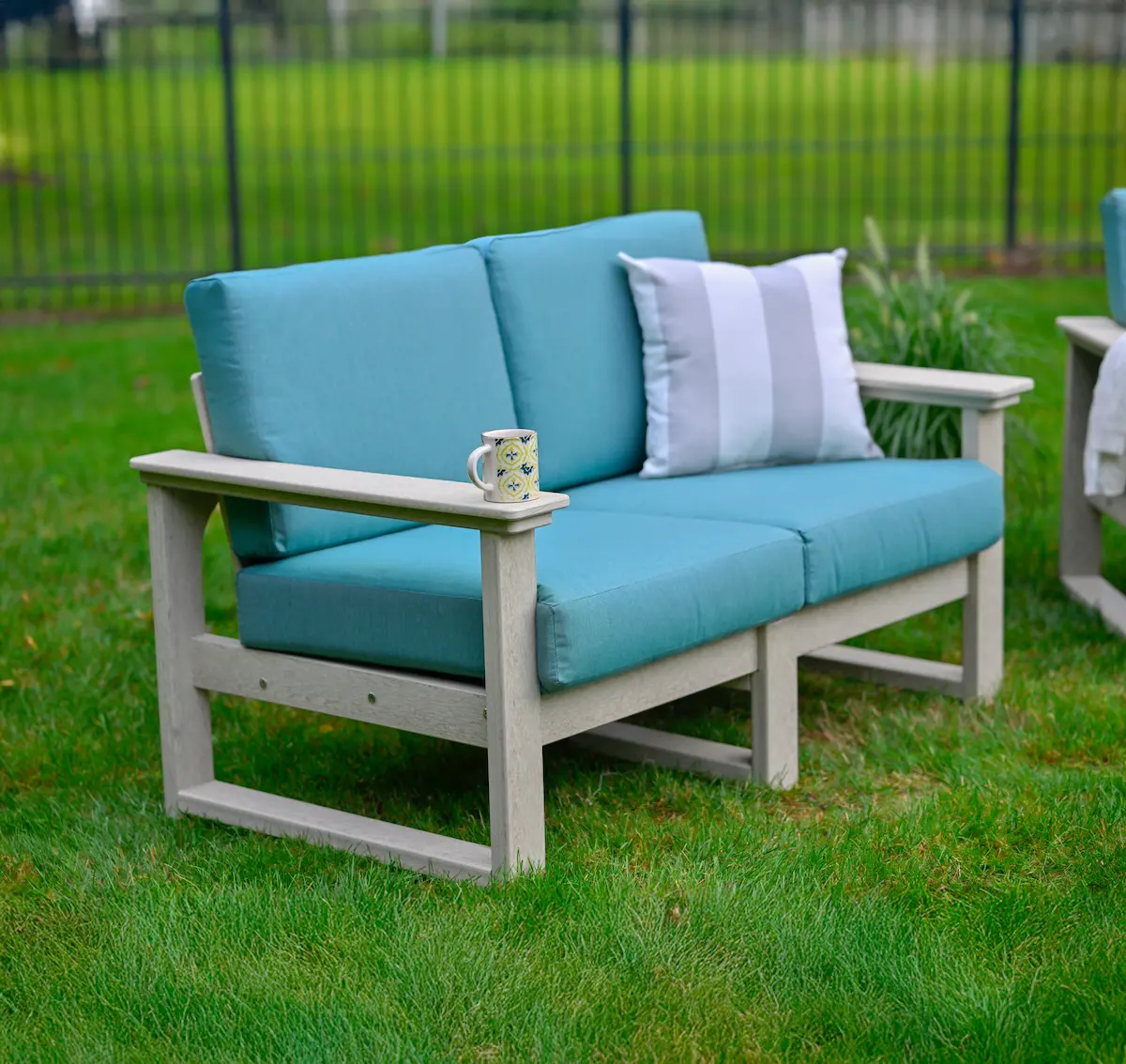 A blue and grey lanai deep seating loveseat with turquoise cushions and a cup holder on the arm. Includes a decorative striped pillow and a mug, positioned on green grass against a dark fence.