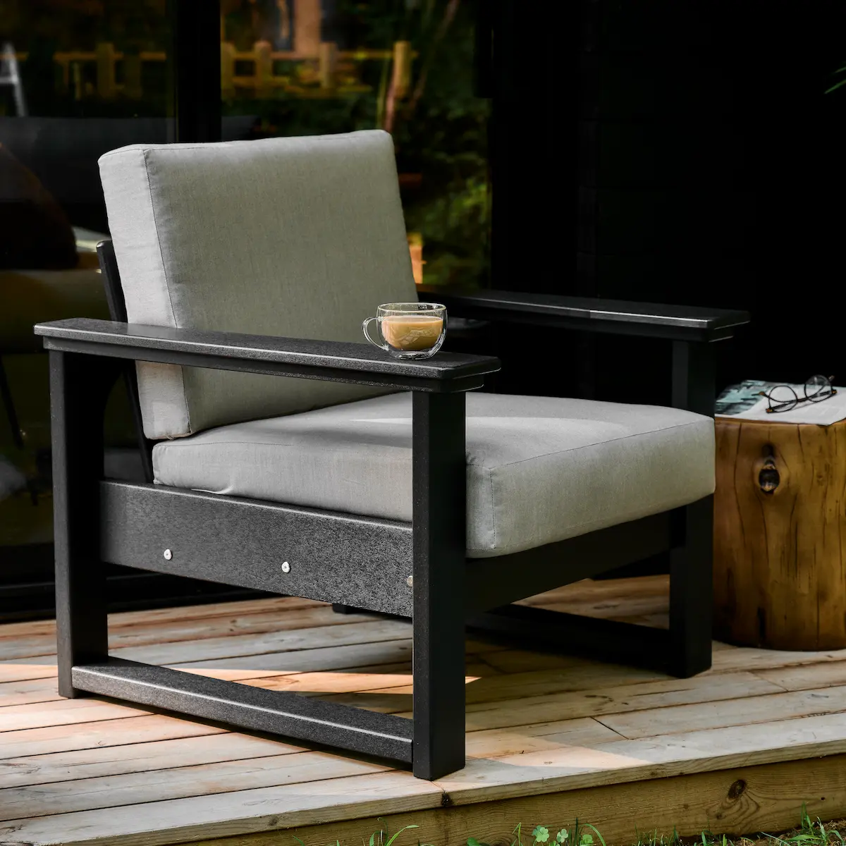 A black and grey lanai deep seating chair with light gray cushions and a glass coffee cup resting on the armrest. Includes a rustic log side table with a magazine and reading glasses, positioned on a wooden deck outdoors.